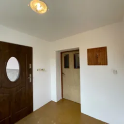 Wnętrze po odświeżeniu: białe ściany, brązowa podłoga, dwoje drzwi - ciemne z owalnym okienkiem i jasne drewnopodobne, oraz mała drewniana szafka na ścianie.