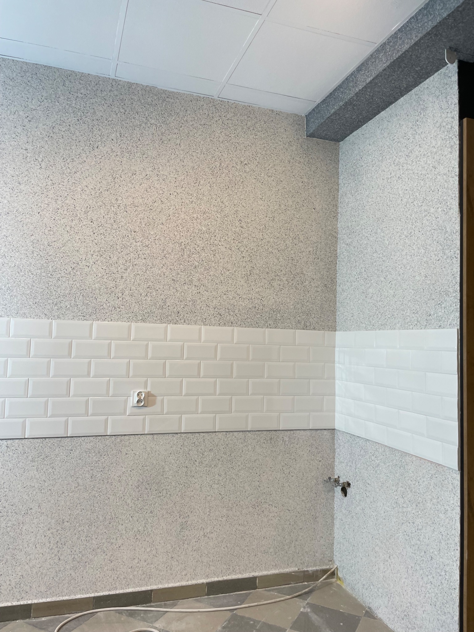 Fragment ściany z dekoracyjną strukturą i białymi, prostokątnymi płytkami ceramicznymi ułożonymi w pasie, widoczny kontakt elektryczny i fragment instalacji wodnej