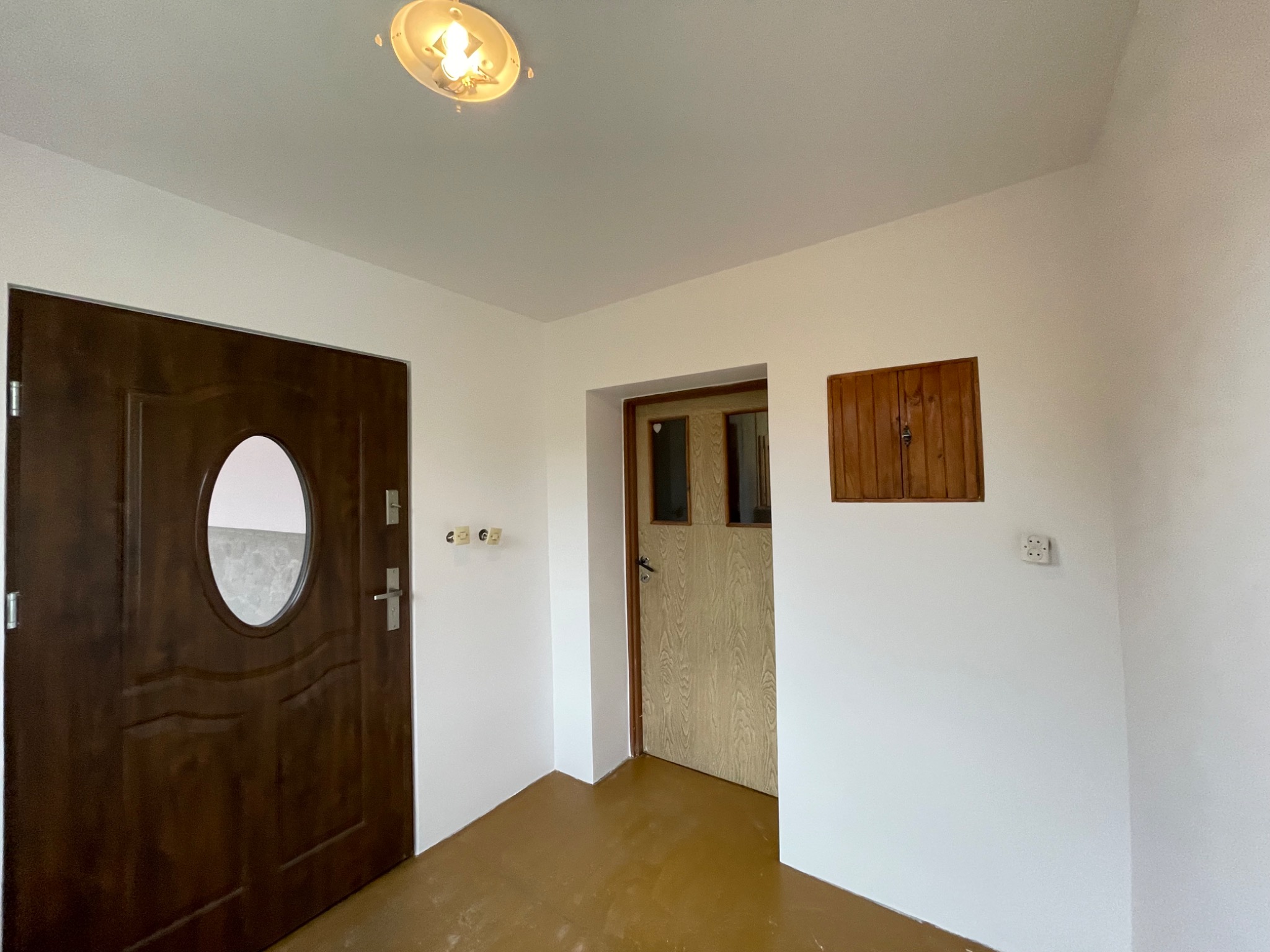 Wnętrze po odświeżeniu: białe ściany, brązowa podłoga, dwoje drzwi - ciemne z owalnym okienkiem i jasne drewnopodobne, oraz mała drewniana szafka na ścianie.