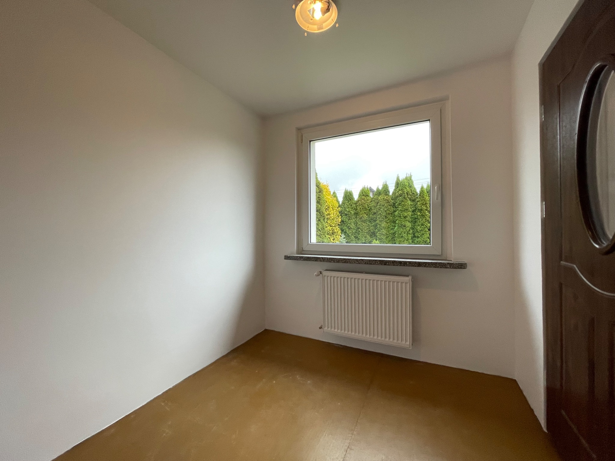 Pusty pokój po remoncie, z białymi ścianami, brązową podłogą zakrytą folią, białym grzejnikiem pod oknem i ciemnymi drzwiami z okrągłym okienkiem, widok na zielone tuje za oknem.