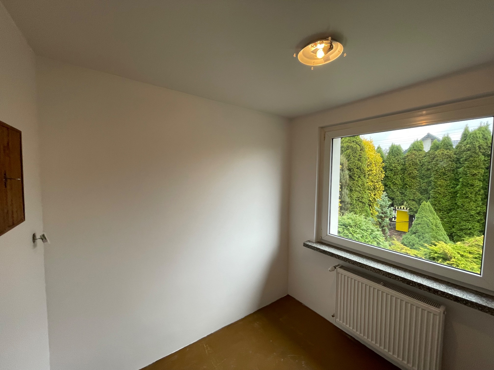 Pusty pokój po remoncie, ściany pomalowane na biało, brązowa podłoga, grzejnik pod oknem z widokiem na zielony ogród, prosta lampa sufitowa.
