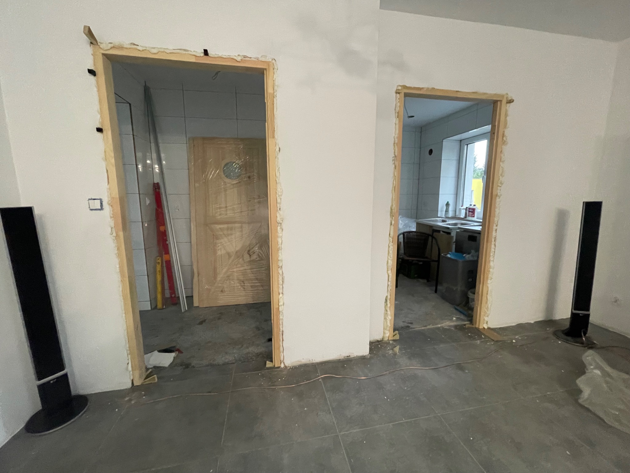 Przygotowane ościeżnice drzwiowe z pianką montażową, widok na łazienkę z płytkami i kuchnię z oknem, szare płytki podłogowe, kolumny głośnikowe w tle.