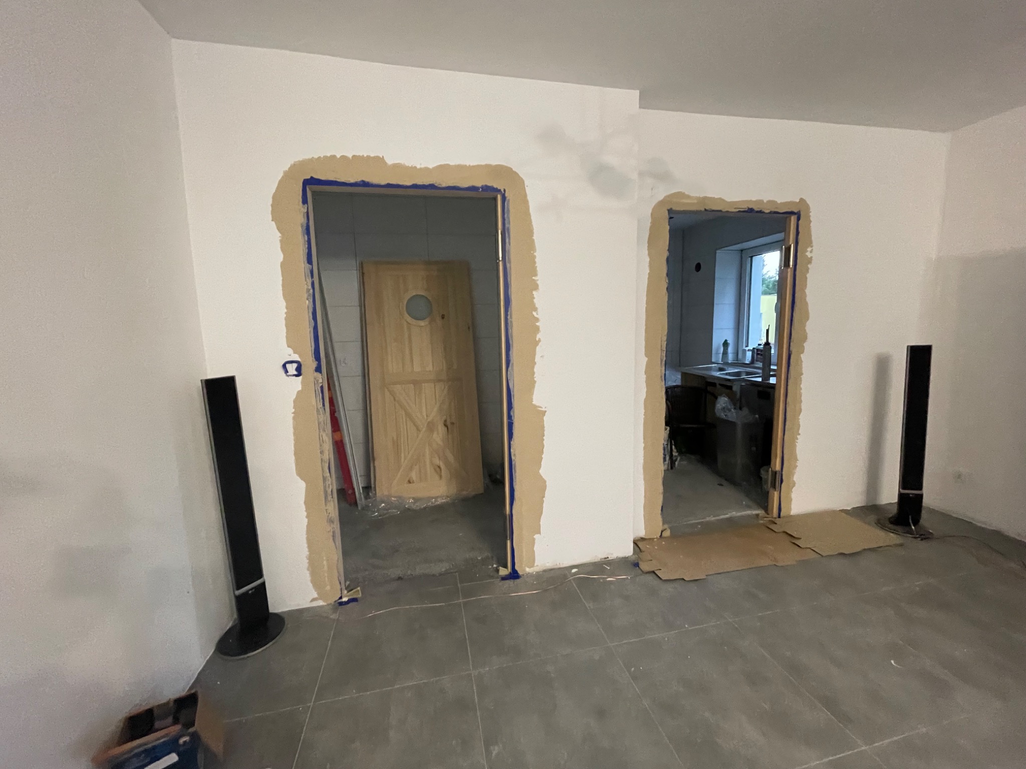 Przygotowane do montażu ościeżnice w dwóch otworach drzwiowych, zabezpieczone taśmą malarską, z widocznymi głośnikami podłogowymi i fragmentem podłogi z szarych płytek.