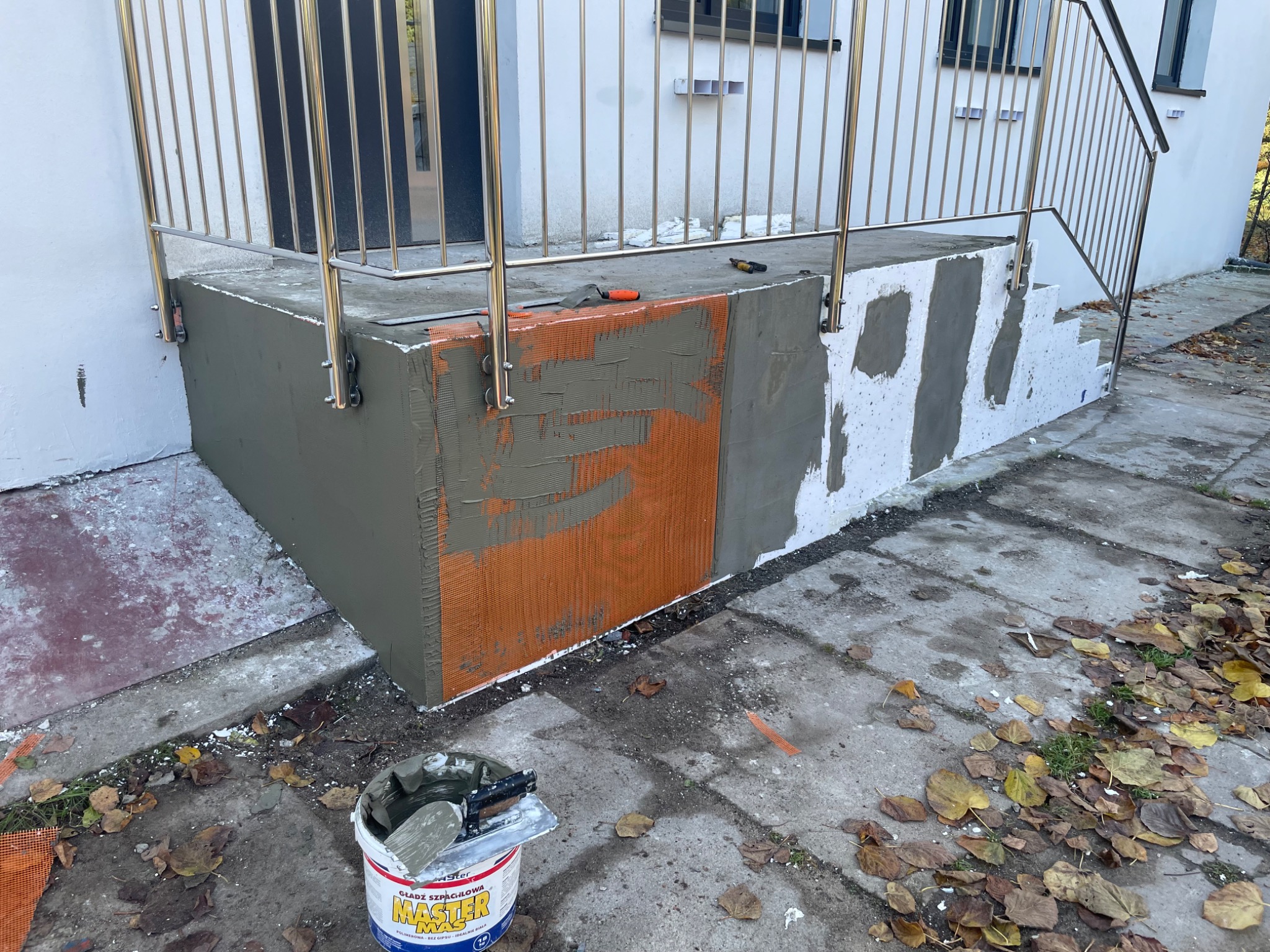 Wykończenie betonowych schodów wejściowych z metalową barierką, nałożona siatka i częściowo szara warstwa wyrównująca, widoczne narzędzia i wiadro z masą szpachlową.