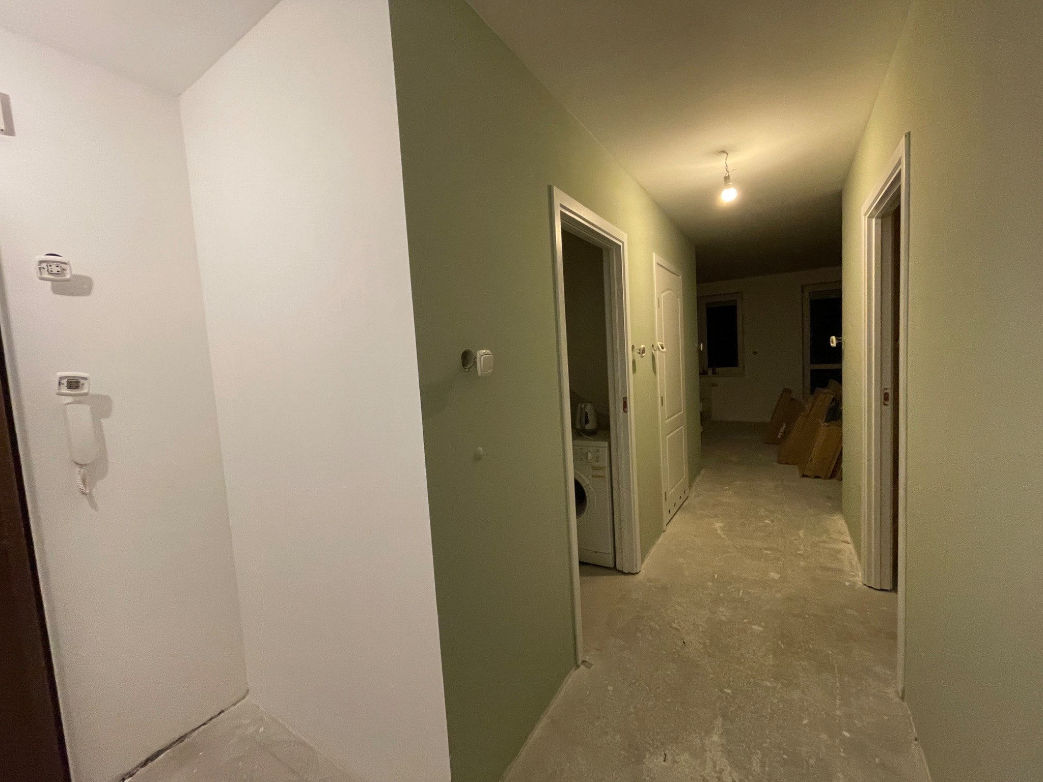 Korytarz w trakcie remontu z świeżo pomalowanymi ścianami na biało i oliwkowo, surową wylewką na podłodze, widoczną pralką w otwartym pomieszczeniu i stertą paneli pod ścianą w głębi.