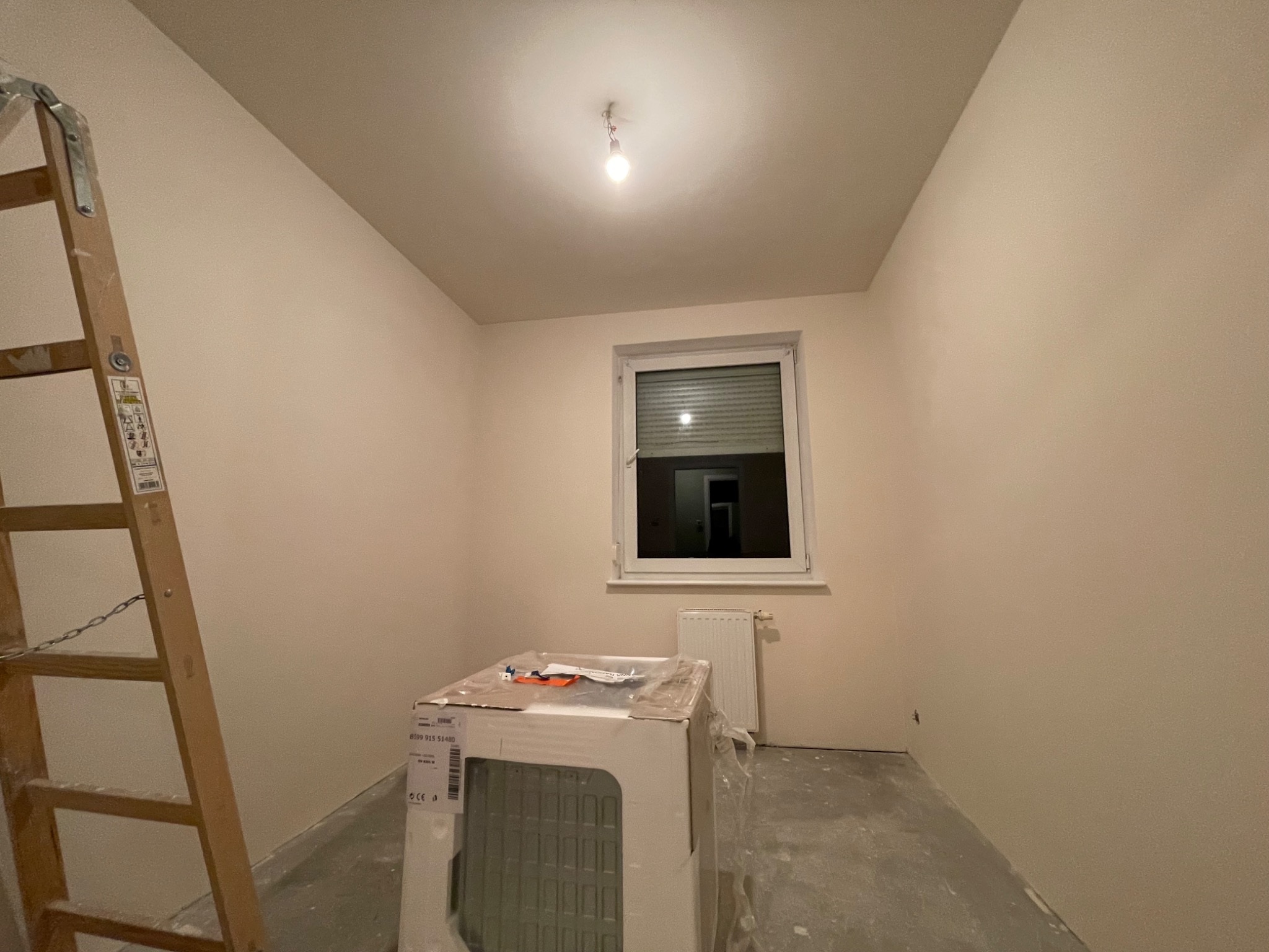 Pokój w trakcie remontu: świeżo pomalowane ściany w odcieniach beżu, drabina drewniana oparta o ścianę, okno z roletami, grzejnik i zapakowany w karton nowy grzejnik.