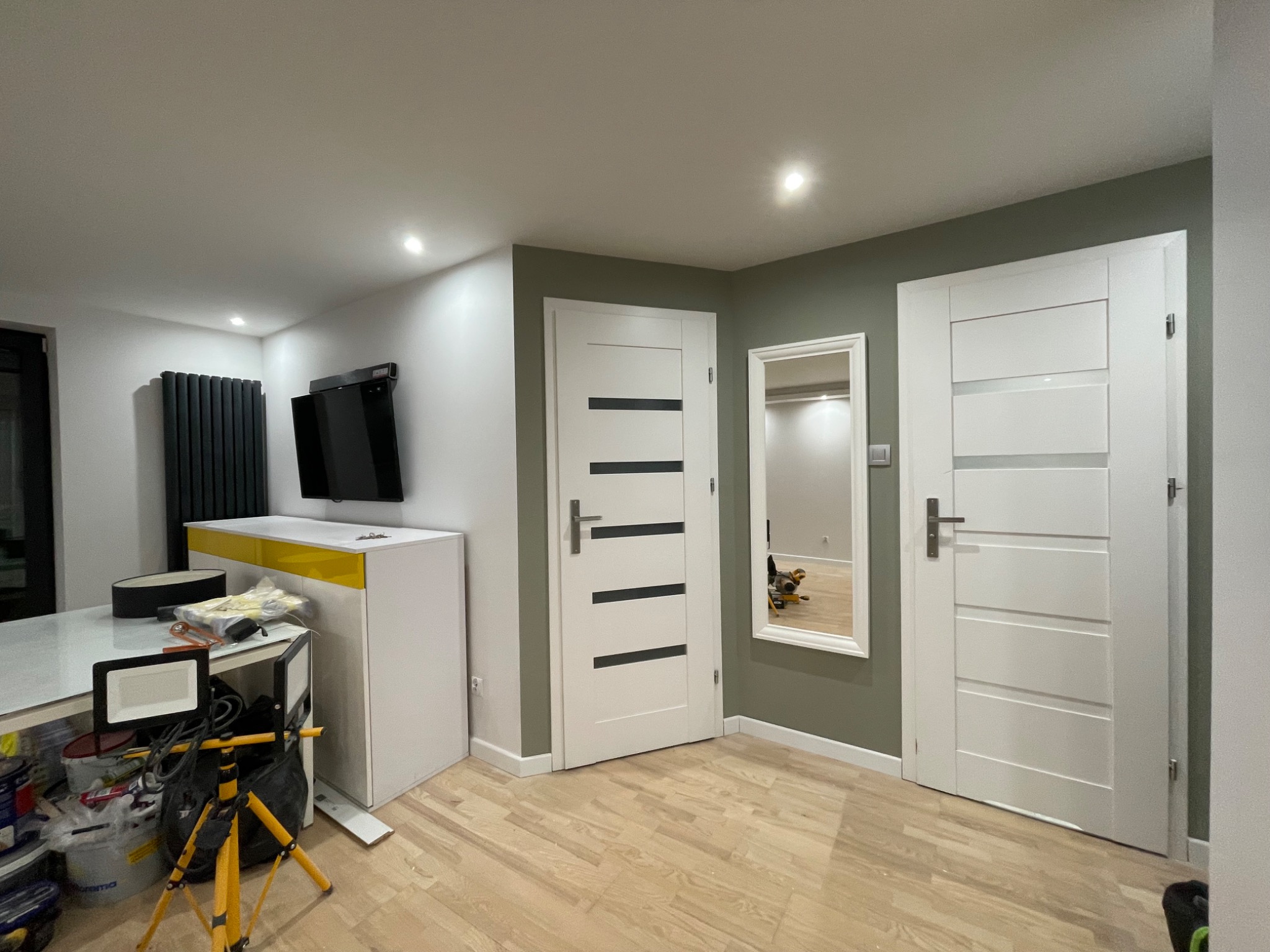 Wnętrze po remoncie z dwiema białymi, nowoczesnymi drzwiami z poziomymi, czarnymi paskami, jasną drewnianą podłogą i ścianą pomalowaną na oliwkowy kolor; w lustrze odbija się fragment pomieszczenia...