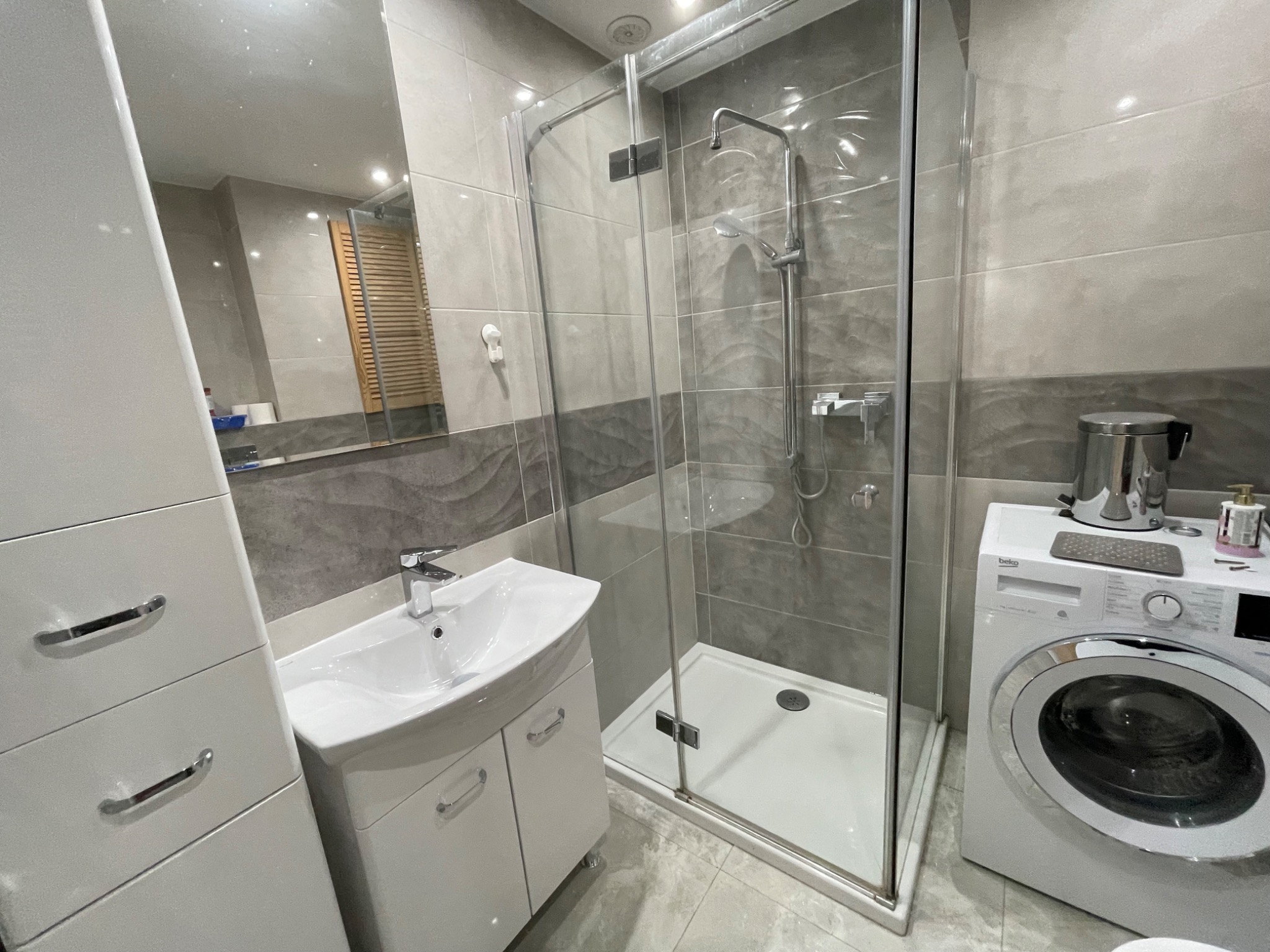 Nowoczesna łazienka z kabiną prysznicową, umywalką z szafką, wysoką szafką i pralką, wykończona szarymi płytkami w dwóch odcieniach, oświetlona górnym światłem.