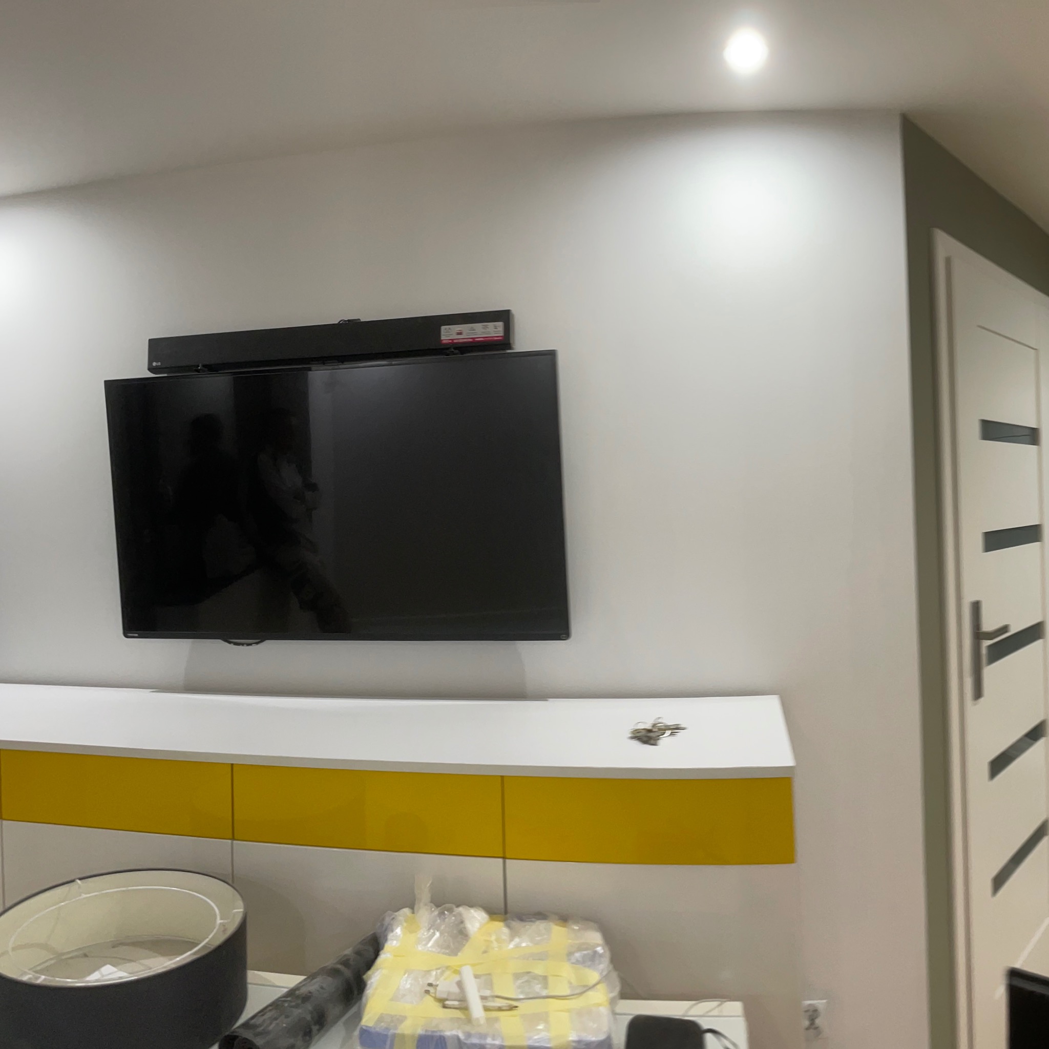 Telewizor zamontowany na ścianie nad biało-żółtą szafką, widoczna również lampa i zapakowane materiały budowlane.