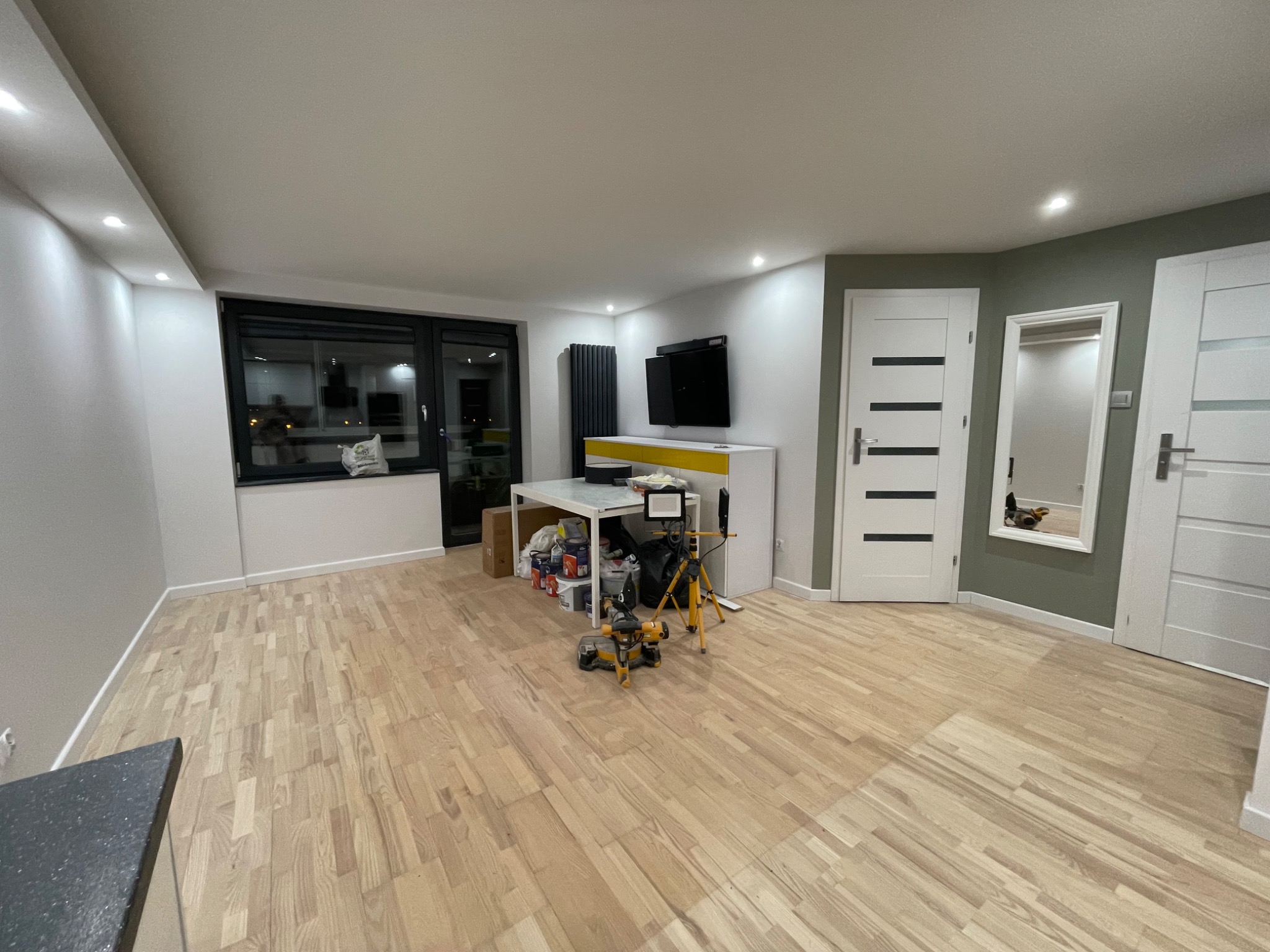 Jasny parkiet ułożony w przestronnym salonie, widoczna piła do cięcia paneli na tle białego stołu z farbami i narzędziami, drzwi wewnętrzne po prawej.