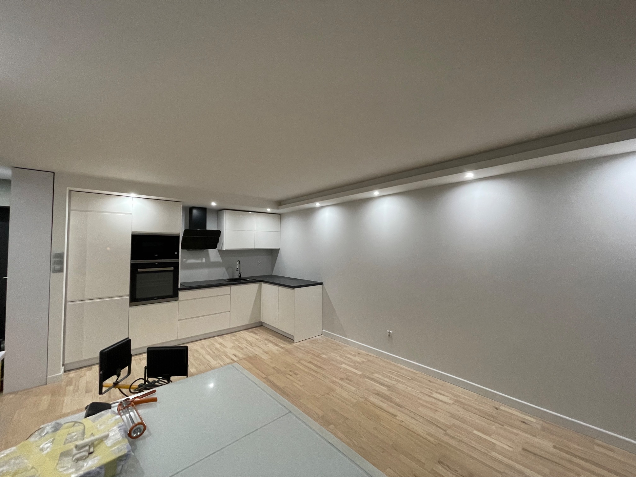 Nowoczesna, jasna kuchnia z białymi szafkami i drewnianą podłogą, w trakcie remontu widoczne narzędzia i zabezpieczenia na blacie stołu.