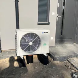 Biała jednostka zewnętrzna klimatyzacji marki York, zamontowana na czarnych wspornikach przy ścianie budynku z szarą stolarką okienną i drzwiami wejściowymi. Widoczny fragment trawnika i kostki...