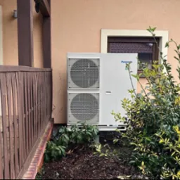 Zewnętrzna jednostka pompy ciepła Panasonic zamontowana przy ścianie domu, częściowo zasłonięta przez krzew z żółto-zielonymi liśćmi, widoczna drewniana balustrada tarasu.