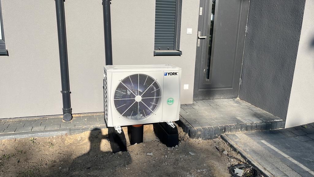 Biała jednostka zewnętrzna klimatyzacji marki York, zamontowana na czarnych wspornikach przy ścianie budynku z szarą stolarką okienną i drzwiami wejściowymi. Widoczny fragment trawnika i kostki...