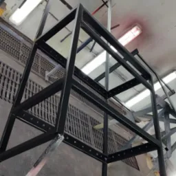 Czarna, metalowa konstrukcja szkieletowa w trakcie malowania, widoczna z dołu w warsztacie z kratami w podłodze i oświetleniem sufitowym.