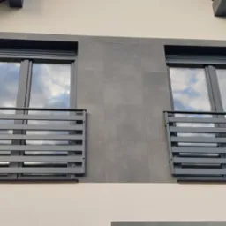 Dwa okna z szarymi roletami i poziomymi, metalowymi balustradami, zamontowane na elewacji domu z szarymi panelami.