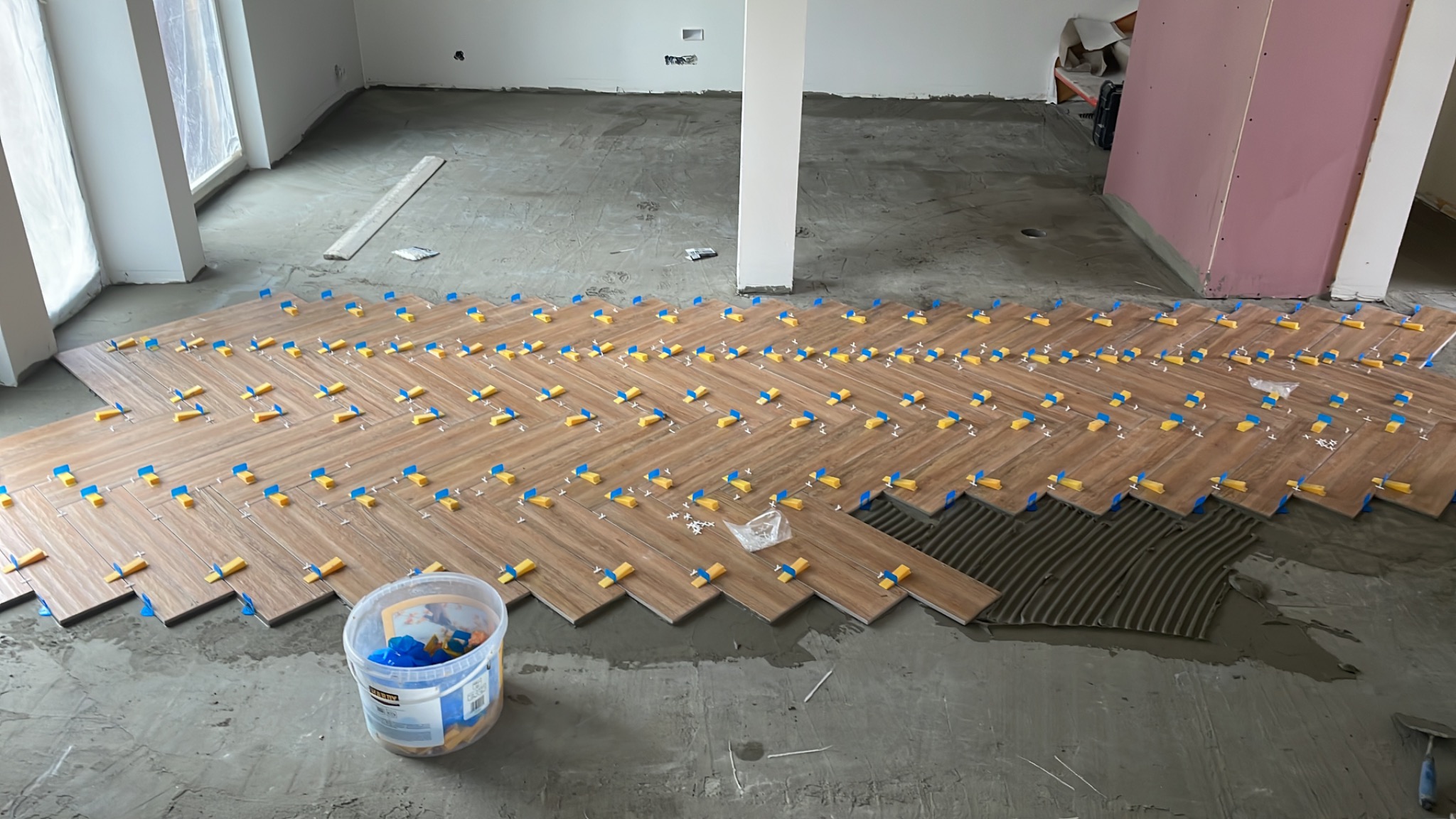 Układanie drewnopodobnej podłogi w jodełkę z użyciem plastikowych krzyżyków dystansowych, widoczny kubeł z krzyżykami i fragment przygotowanego podłoża z nałożonym klejem.