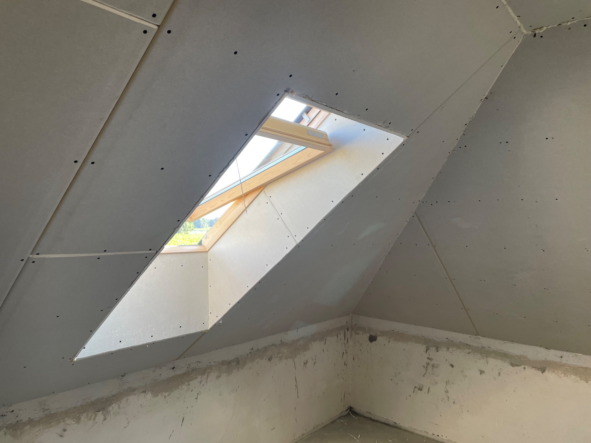 Poddasze w trakcie wykańczania: zamontowane płyty gipsowo-kartonowe na skosach dachu z oknem połaciowym, ściany poniżej przygotowane do dalszych prac.