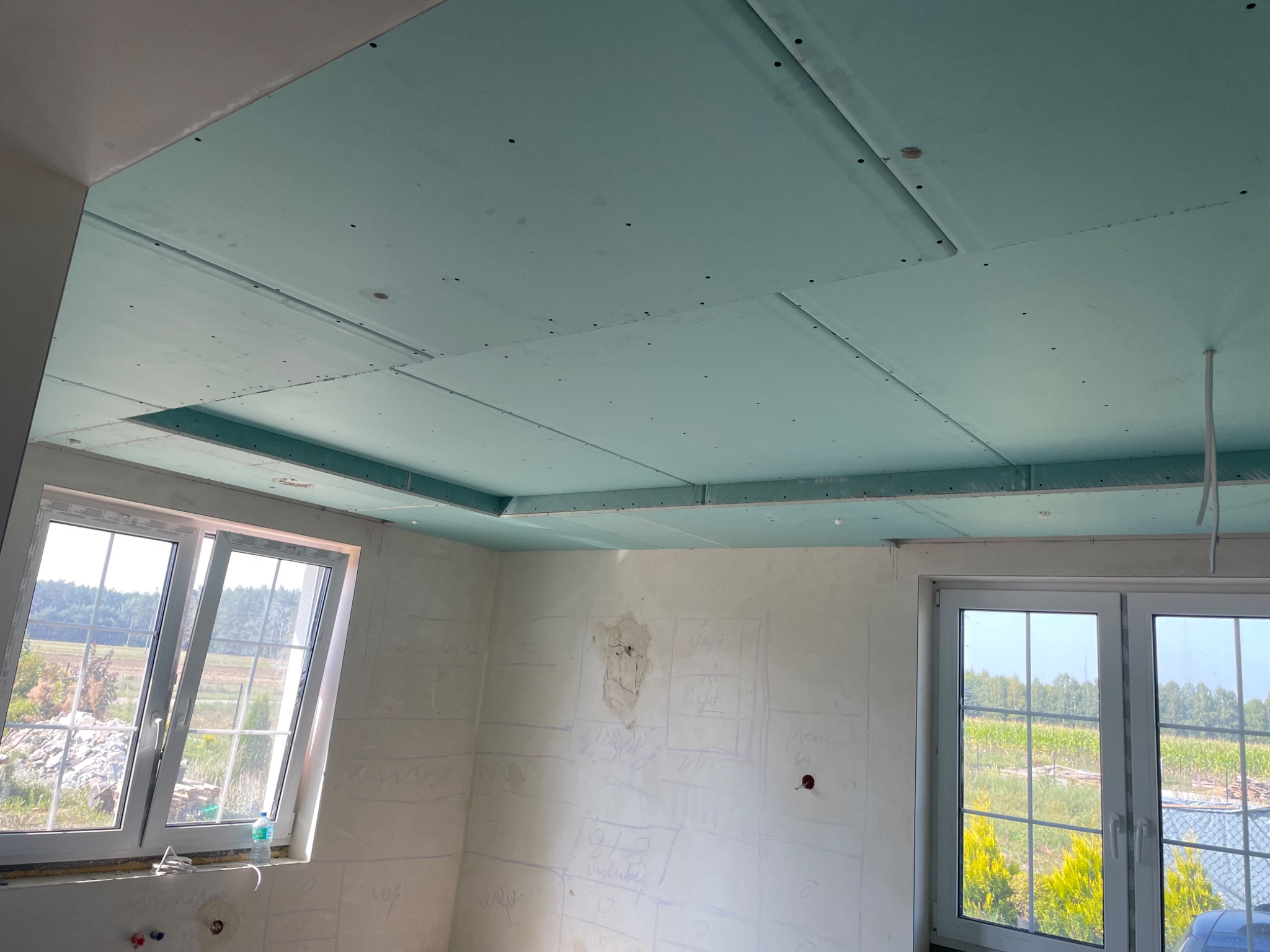 Niebieskie płyty kartonowo-gipsowe zamontowane na suficie podwieszanym, ściany z widocznymi szkicami instalacji, okna z widokiem na zewnątrz.