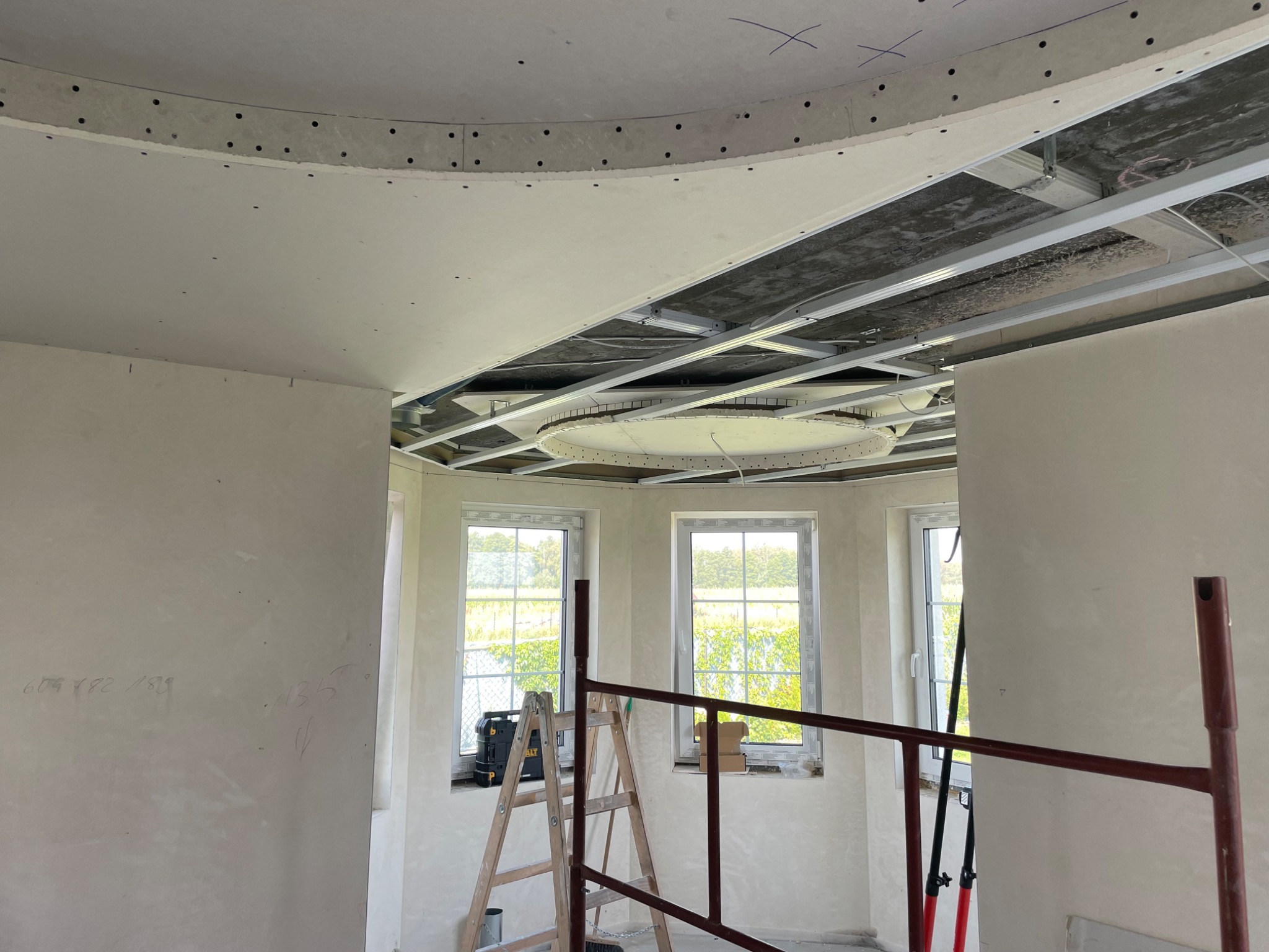 Wnętrze w trakcie prac wykończeniowych: widoczna konstrukcja sufitu podwieszanego z płyt kartonowo-gipsowych o nieregularnym kształcie, częściowo zamontowana, z widocznymi profilami metalowymi...