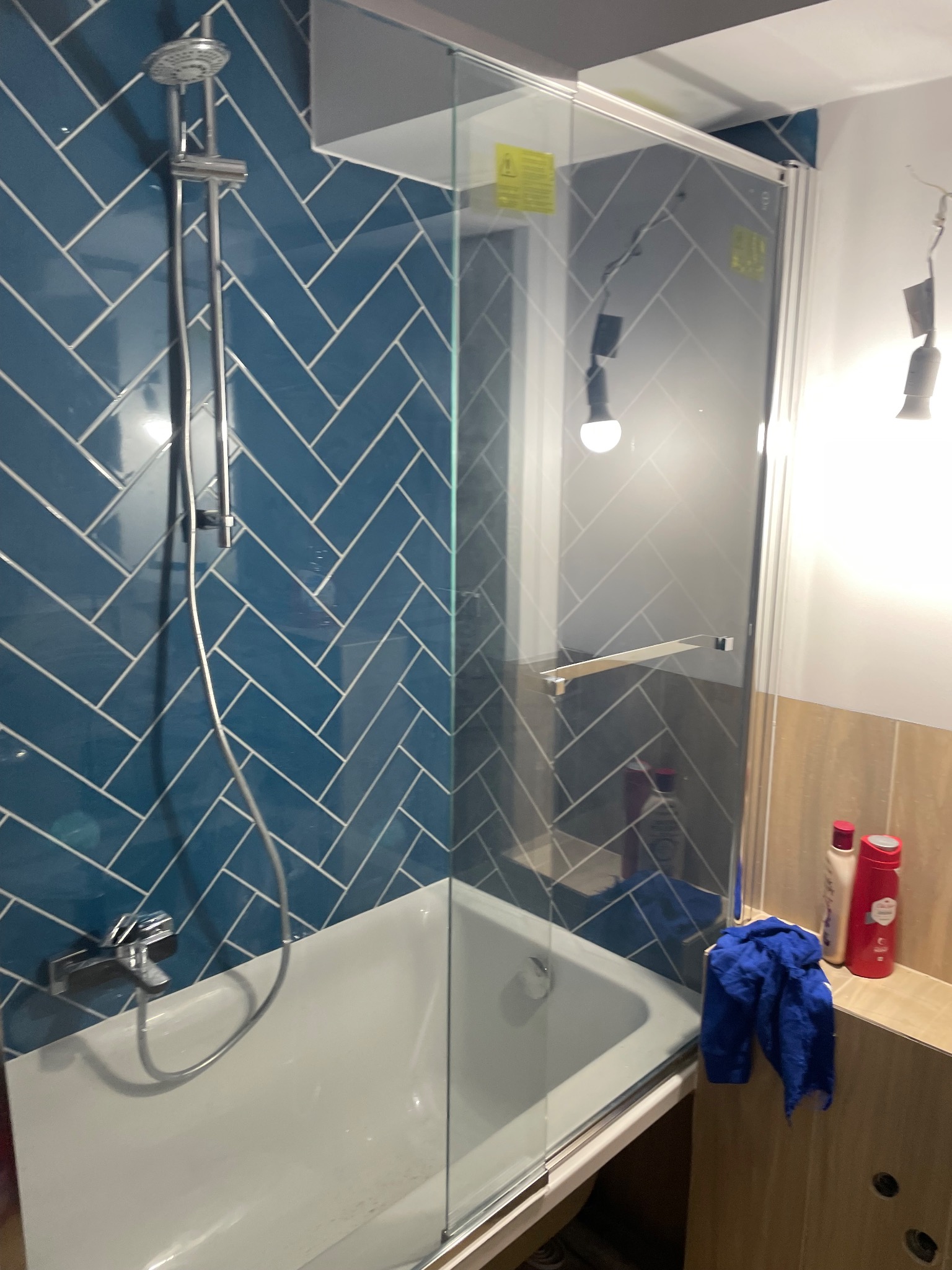 Niebieska łazienka z płytkami ułożonymi w jodełkę, kabina prysznicowa ze szkła hartowanego, bateria wannowa z chromowanym wykończeniem, niebieski ręcznik obok kosmetyków.