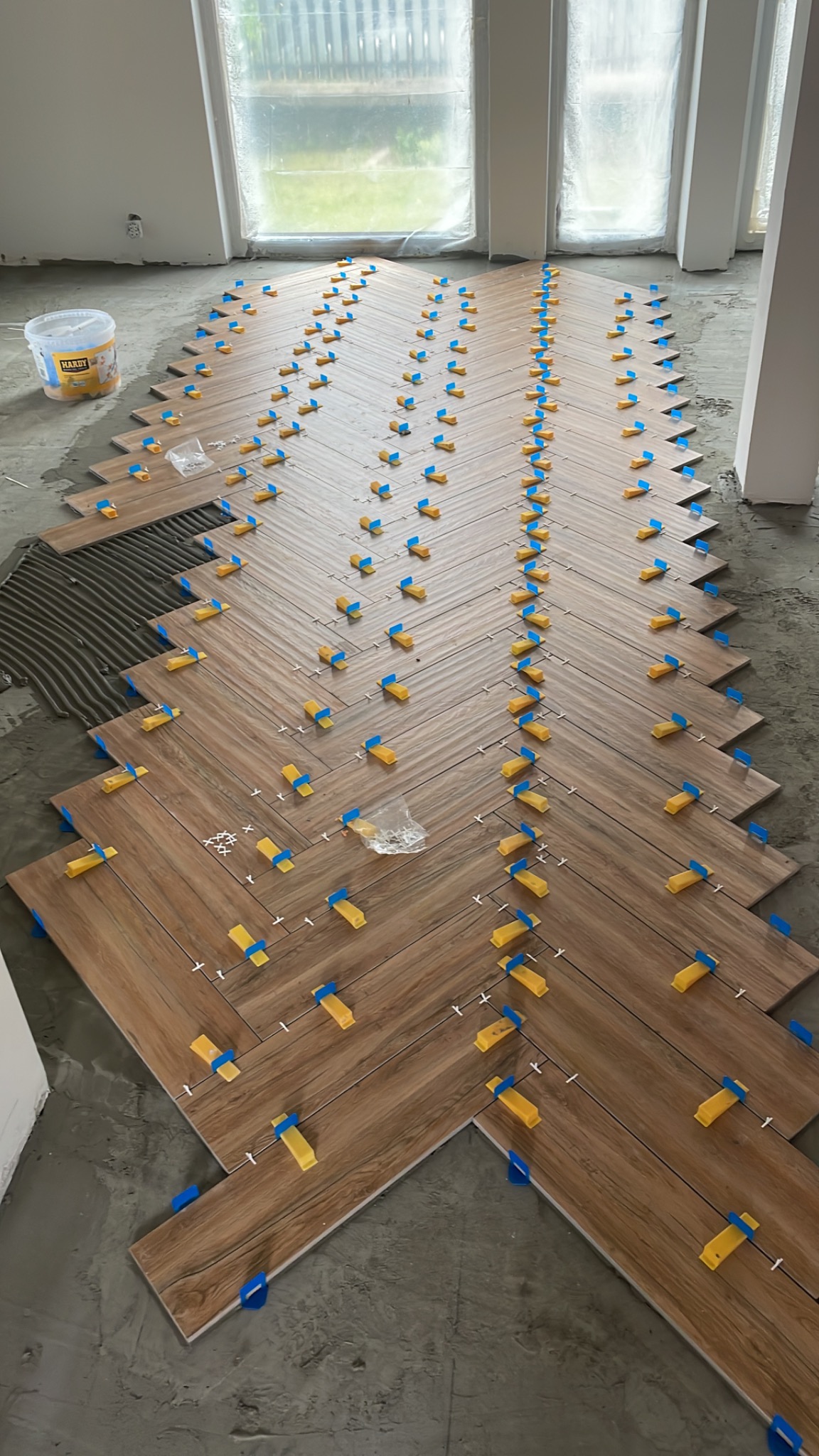 Montaż parkietu w jodełkę z użyciem systemu poziomowania płytek; widoczne brązowe panele, niebieskie kliny i żółte elementy dociskowe, wiadro z klejem w tle.