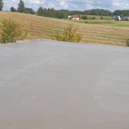 KBB Usługi Ogólnobudowlane - Świeżo wylany, gładki fundament płyty budynku na tle pagórkowatego krajobrazu z zabudowaniami i zielenią.