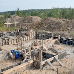 KBB Usługi Ogólnobudowlane - Zalewanie betonem szalunków fundamentowych zbrojonych stalą, widoczne niebieskie rury instalacyjne, ziemia usypana w tle.