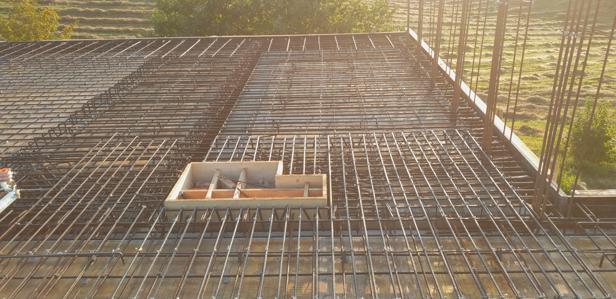 Widok z góry na zbrojenie stropu z siatki stalowej, z drewnianym szalunkiem w centralnej części, w tle widoczne łąki i drzewa w porannym świetle.