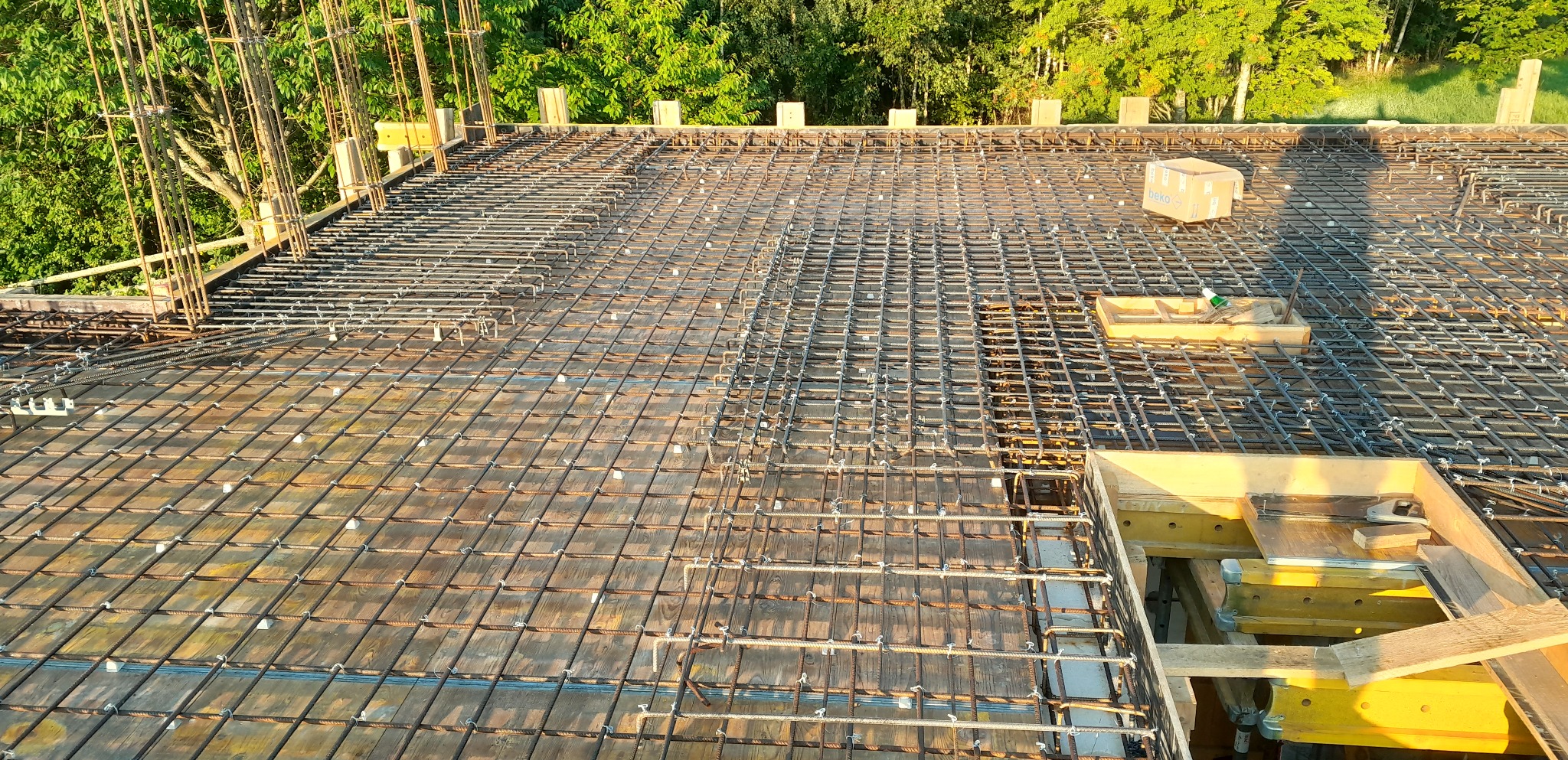 Siatka zbrojeniowa na deskowaniu stropu, widoczne elementy mocujące, drewniane szalunki oraz kartonowe pudło Beko w tle.