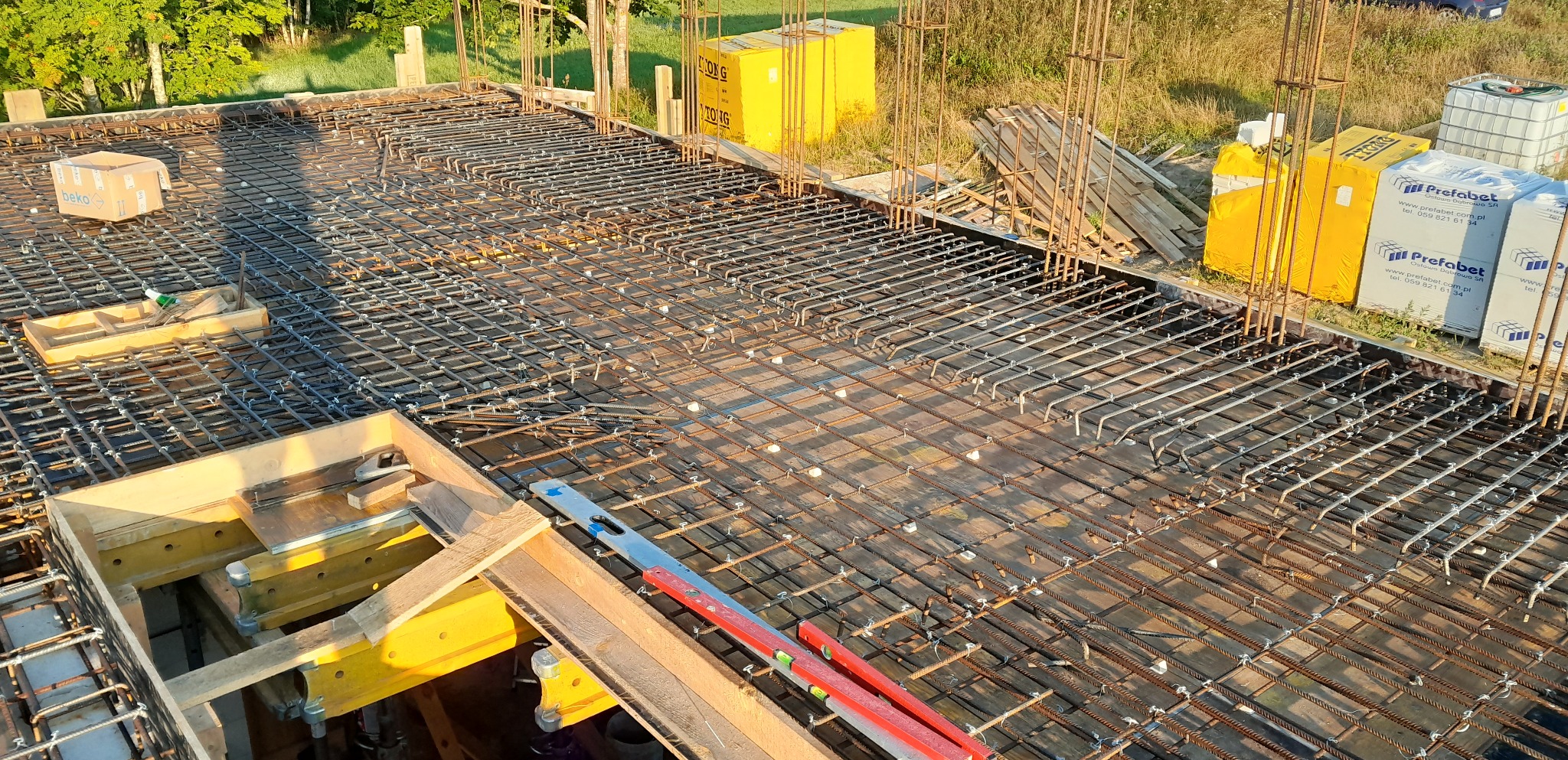 Siatka zbrojeniowa stropu z widocznymi prętami, poziomica i opakowaniami materiałów budowlanych w tle, częściowo w drewnianej ramie szalunkowej.