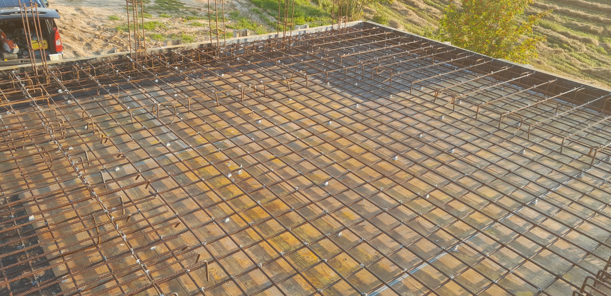 Siatka zbrojeniowa ułożona na szalunku z desek przed zalaniem betonem, widok z góry, z pojazdem i zielenią w tle.