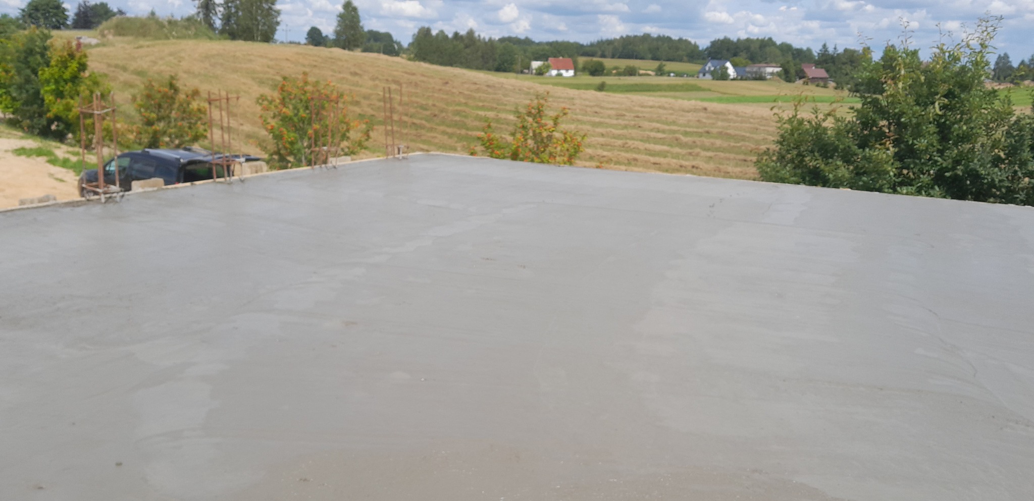 Świeżo wylany, gładki fundament płyty budynku na tle pagórkowatego krajobrazu z zabudowaniami i zielenią.