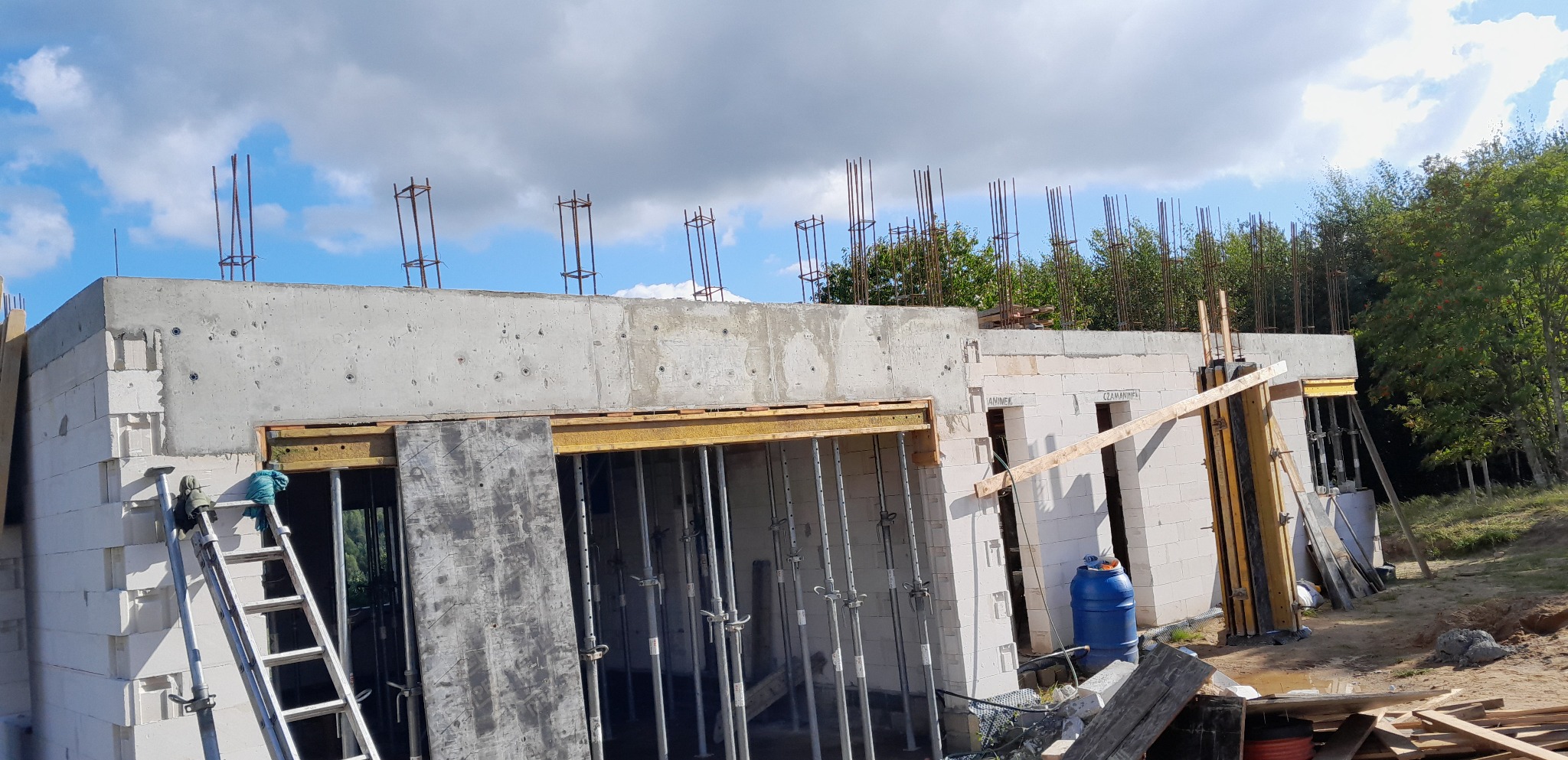 Wznoszenie ścian budynku z betonu komórkowego, widoczne szalunki i zbrojenie, drabina oparta o ścianę, niebieska beczka w tle.