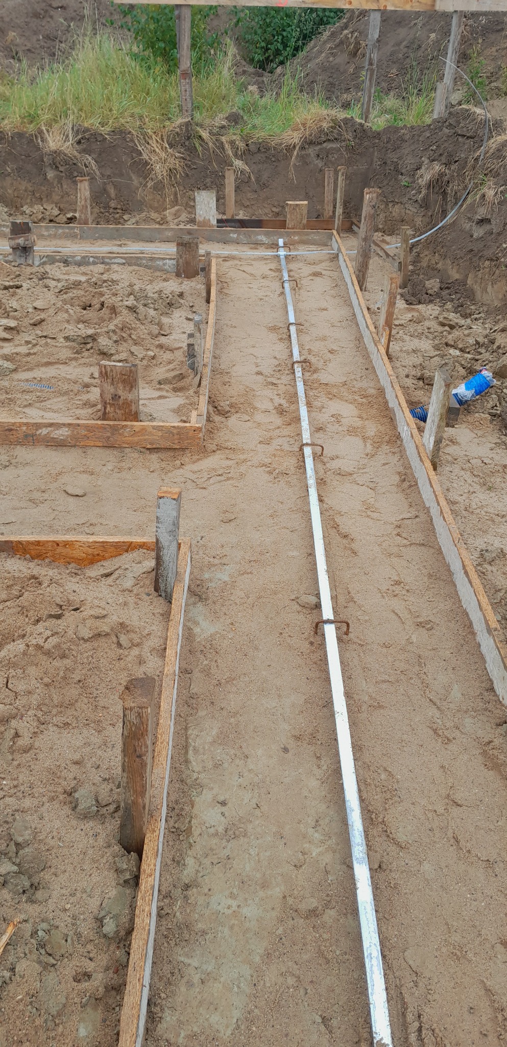 Przygotowanie szalunku pod fundamenty z drewnianych desek i metalowych rurek w wykopie, widoczna ziemia i trawa w tle.