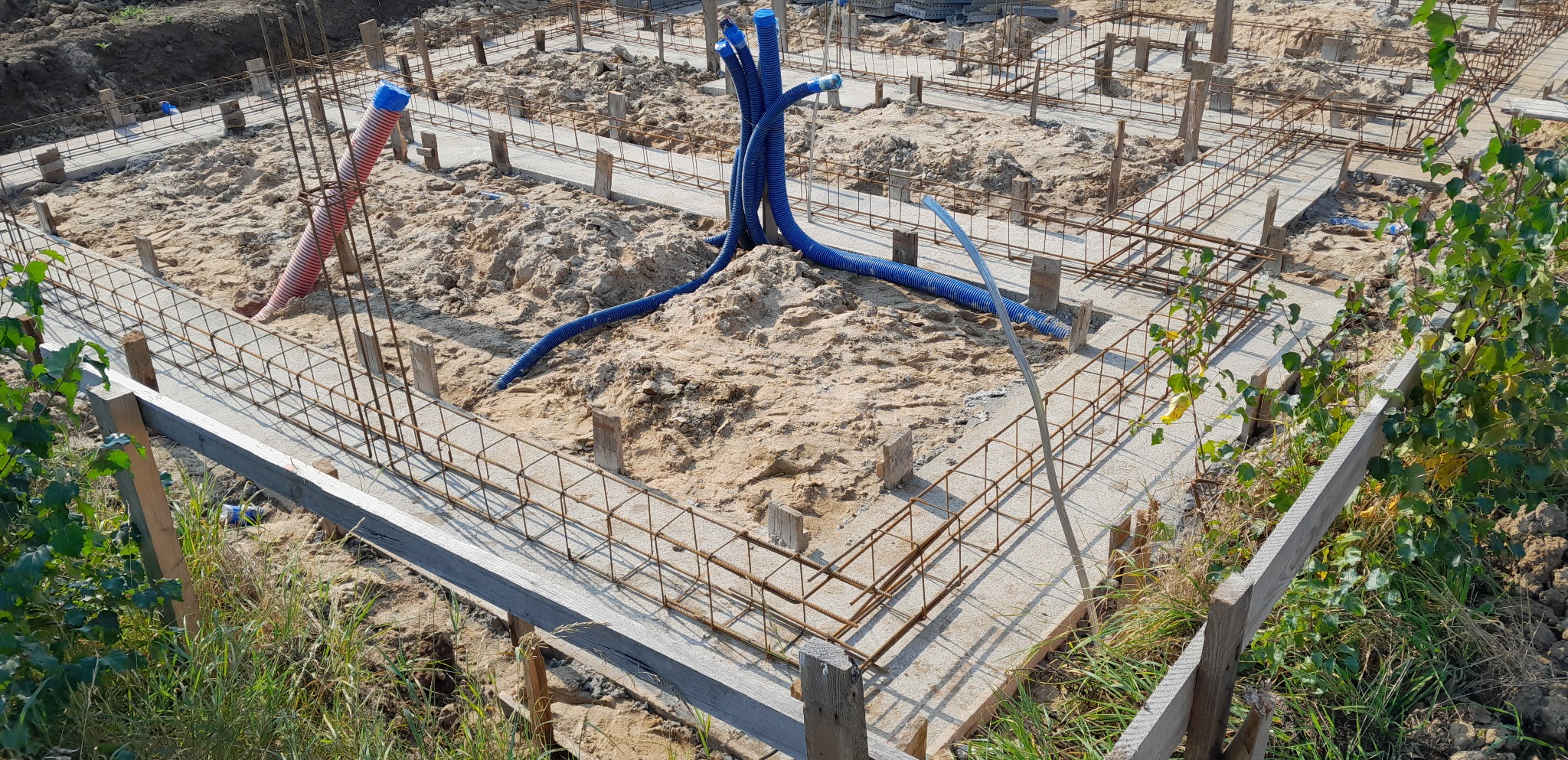 Zalane betonem fundamenty z wystającymi pionowymi prętami zbrojeniowymi, widoczne niebieskie rury instalacyjne i fragment czerwonej rury, teren budowy z piaskiem i fragmentami zieleni.