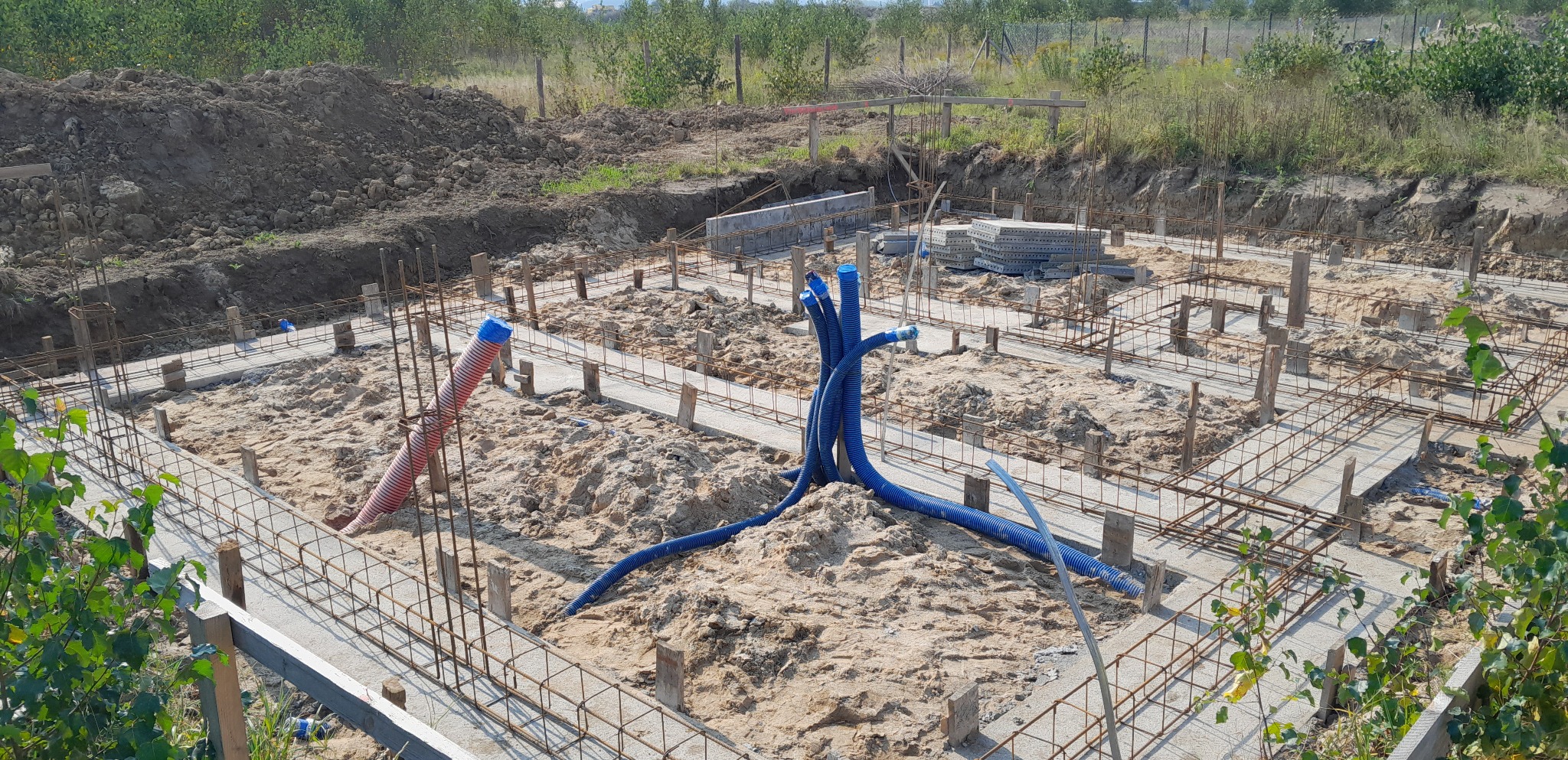 Zbrojone fundamenty w trakcie budowy, widoczne wystające rury instalacyjne w kolorze niebieskim i czerwonym, teren przygotowany pod wylewkę, w tle wykopana ziemia.