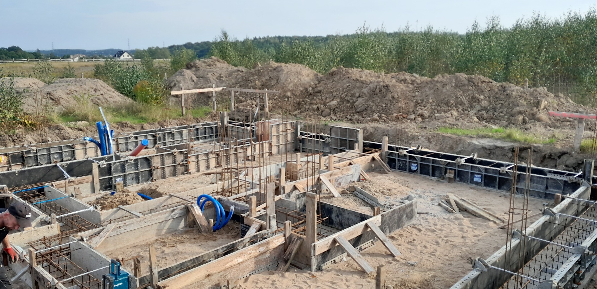 Zalewanie betonem szalunków fundamentowych zbrojonych stalą, widoczne niebieskie rury instalacyjne, ziemia usypana w tle.