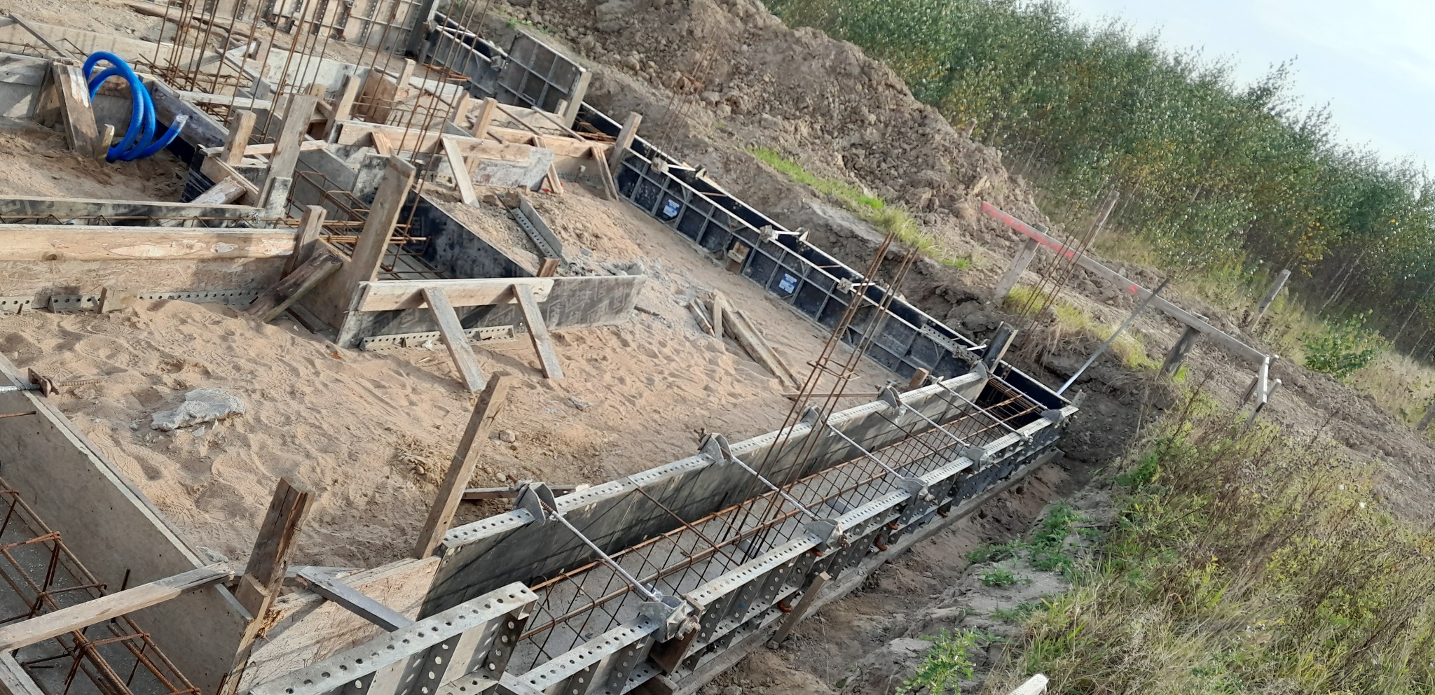 Przygotowanie do wylewki fundamentów: szalunki z metalowymi wzmocnieniami i zbrojenie stalowe w wykopie, widoczne niebieskie rury instalacyjne.