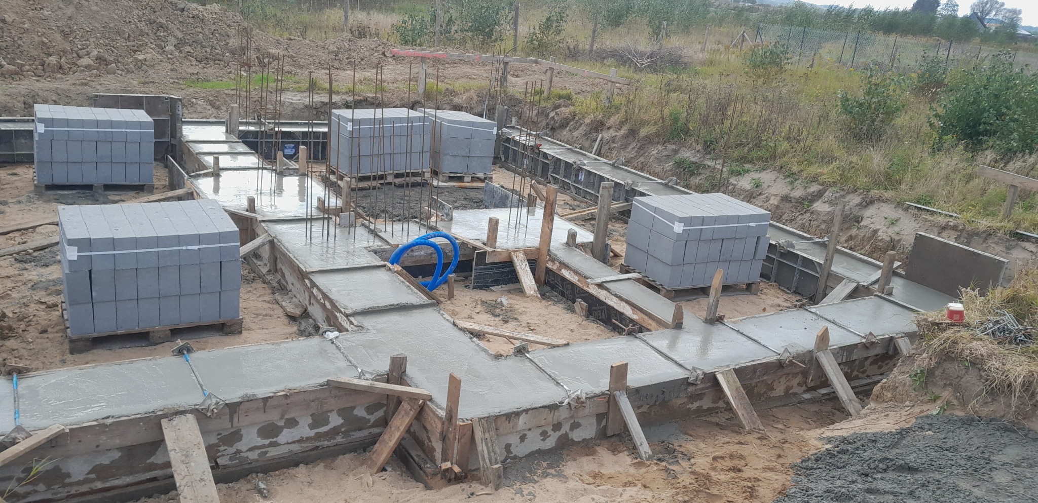Zalane betonem szalunki fundamentowe z wystającymi prętami zbrojeniowymi, obok ułożone bloczki betonowe na paletach i niebieski wąż na budowie w terenie niezabudowanym.