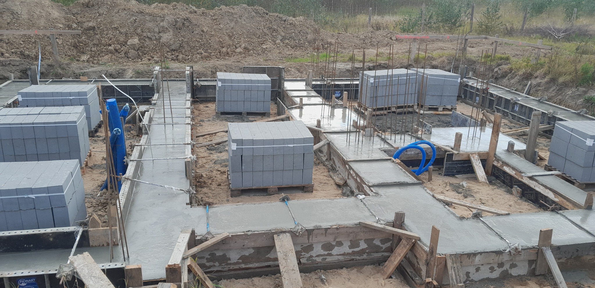 Zalany beton w szalunku pod fundamenty domu z widocznymi zbrojeniami pionowymi i przygotowanymi bloczkami betonowymi. Przez szalunek przechodzą niebieskie rury.