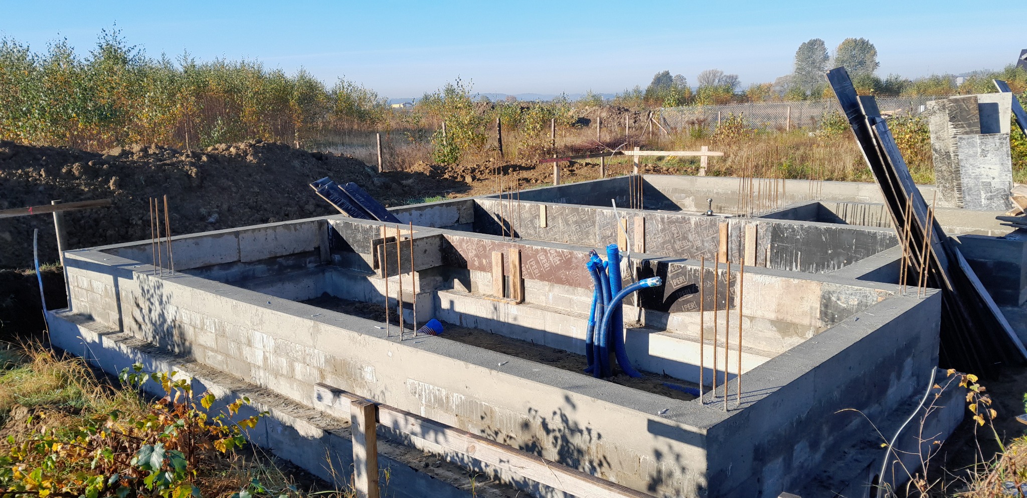 Wylany fundament z bloczków betonowych z wystającymi prętami zbrojeniowymi i niebieskimi rurami instalacyjnymi, widok z góry na tle krajobrazu.
