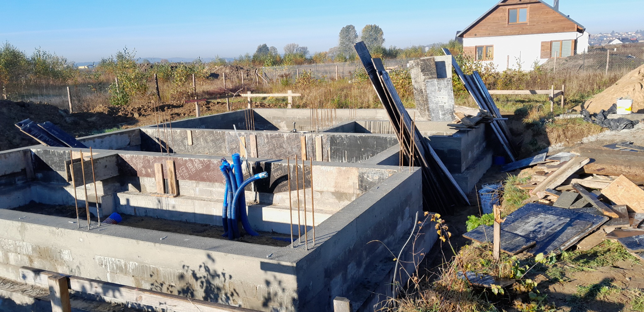 Wylany fundament z wystającymi prętami zbrojeniowymi, widoczne niebieskie rury instalacyjne, w tle dom jednorodzinny w stanie surowym otwartym i krajobraz podmiejski.
