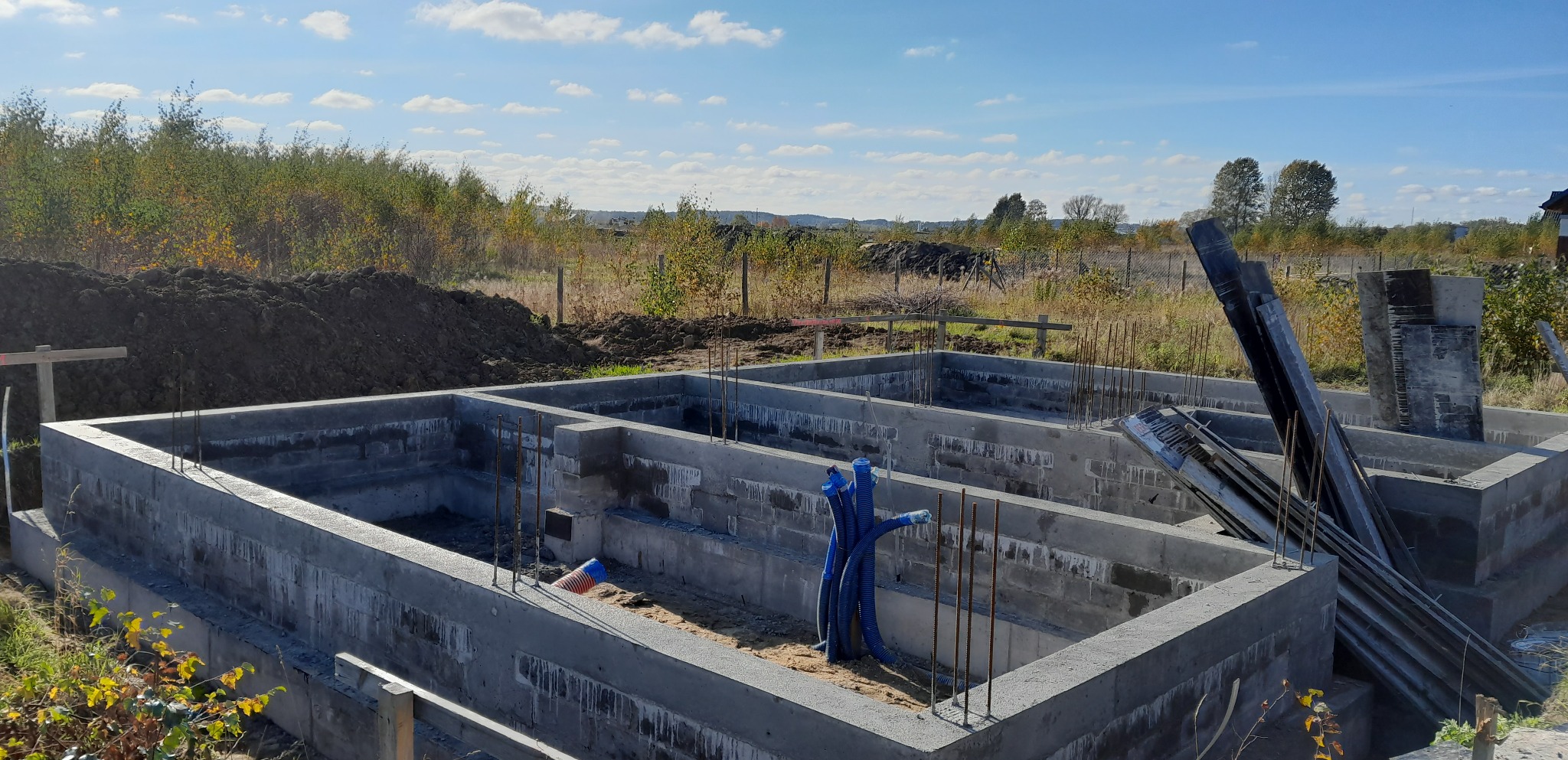 Wylane fundamenty z wystającymi prętami zbrojeniowymi, niebieskie rury instalacyjne wewnątrz, ziemia i drzewa w tle, słoneczny dzień.