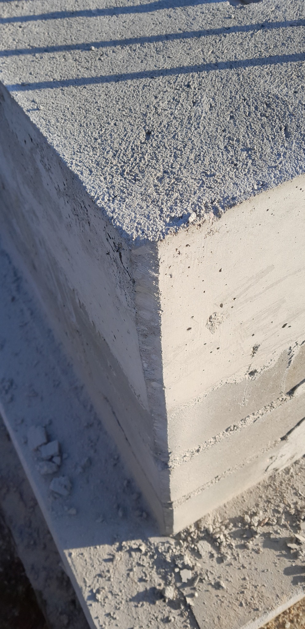 Szczegółowy widok narożnika betonowego fundamentu z widoczną fakturą i warstwami betonu, oświetlonego bocznym światłem słonecznym.