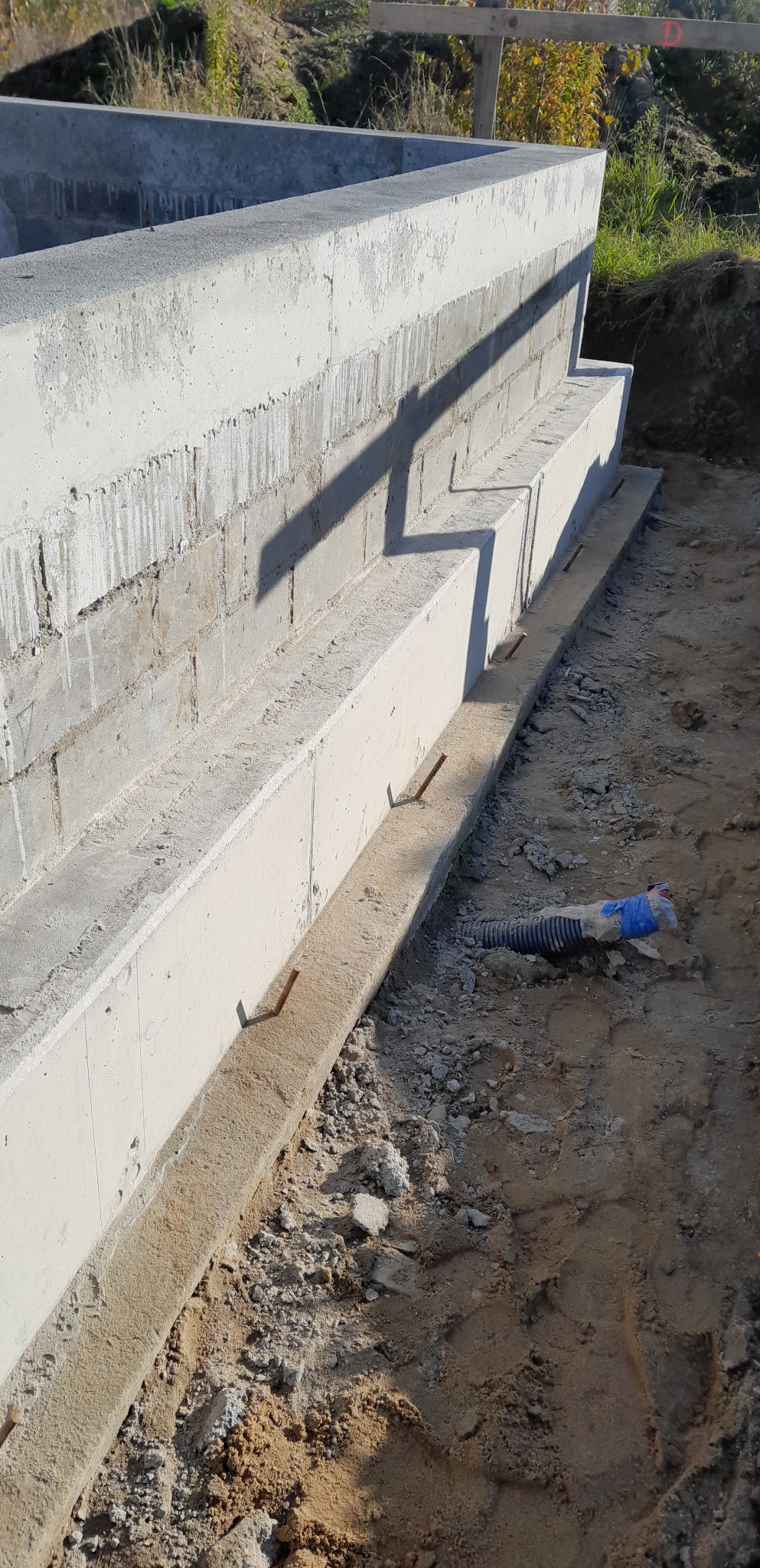 Betonowe ściany fundamentowe z wystającymi prętami zbrojeniowymi, widoczny fragment rury w ziemi, etap budowy.