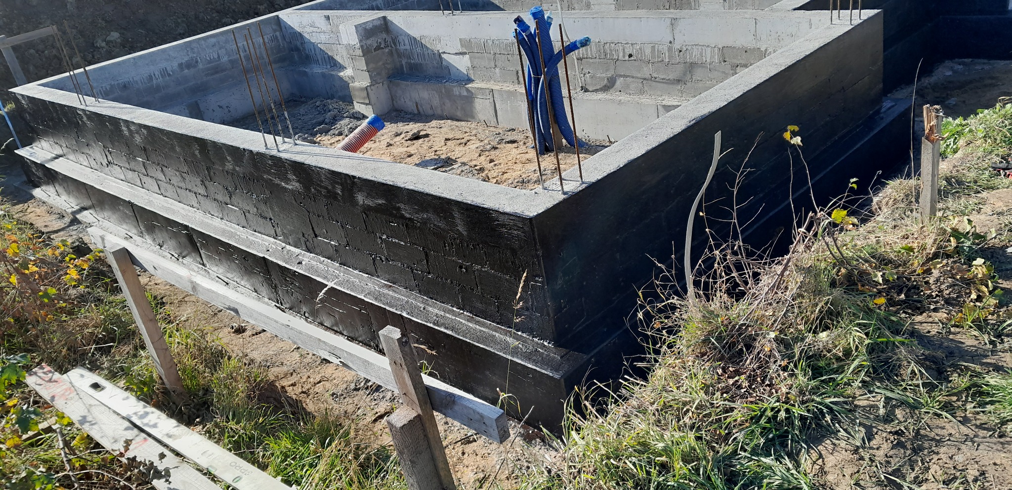 Zabezpieczone hydroizolacją fundamenty z betonu komórkowego, wystające pręty zbrojeniowe, niebieskie rury instalacyjne, ziemia wewnątrz wykopu.