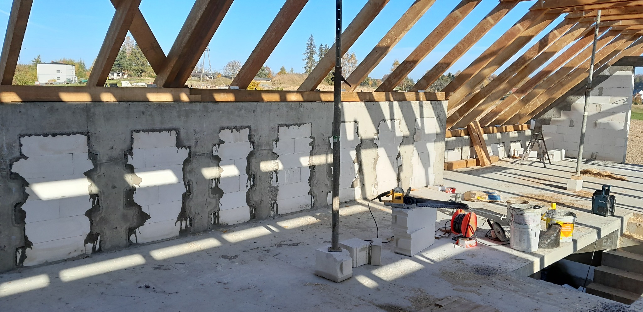 Wnętrze domu w trakcie budowy: ściany z betonu komórkowego z widocznymi połączeniami, drewniana konstrukcja dachu, narzędzia budowlane i materiały leżące na podłodze, wpadające promienie słoneczne.