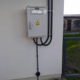 Szara skrzynka elektryczna z symbolem ostrzegawczym zamontowana na elewacji budynku, z widocznymi przewodami w peszlach przymocowanymi do ściany oraz uziemieniem.