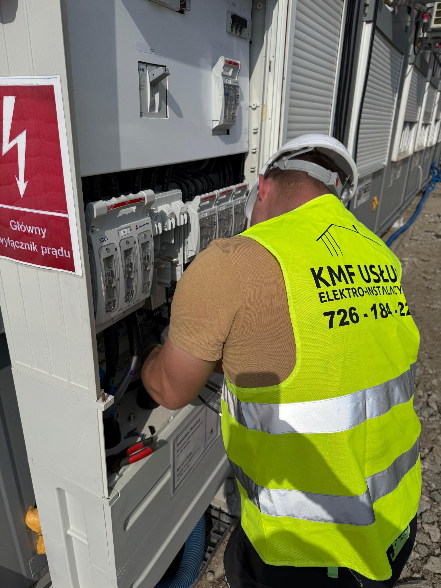 Elektryk w żółtej kamizelce z logo firmy KMF Usługi Elektro-Instalacje, w kasku ochronnym, pracuje przy otwartej skrzynce elektrycznej z bezpiecznikami RBK1 i napisem 'Główny wyłącznik prądu'...
